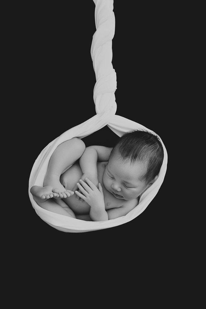 Black and white artistic newborn portrait of a baby curled within white fabric against a dark background, created by a newborn photographer serving Braintree, Essex.