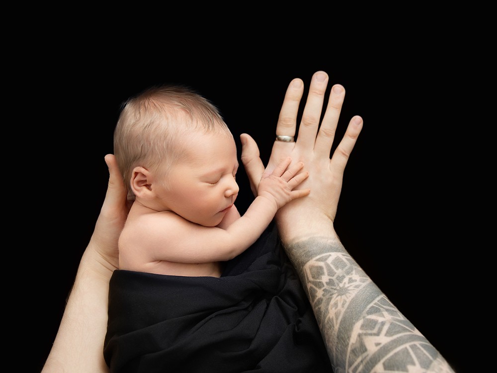 Sleeping newborn baby resting against father's hands with baby's tiny hand against his palm, photographed during an in-home newborn session near Hatfield Peverel, Essex.