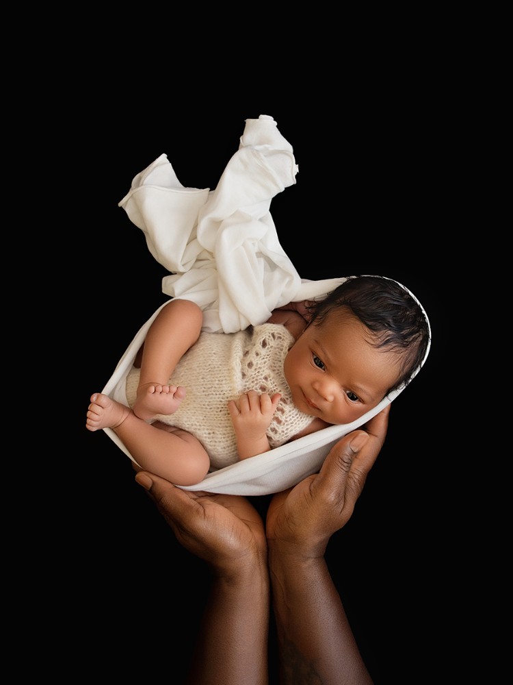Newborn baby gently cradled in a parent's hands with flowing white fabric against a black background, photographed during a bespoke newborn photography session in Witham, Essex.