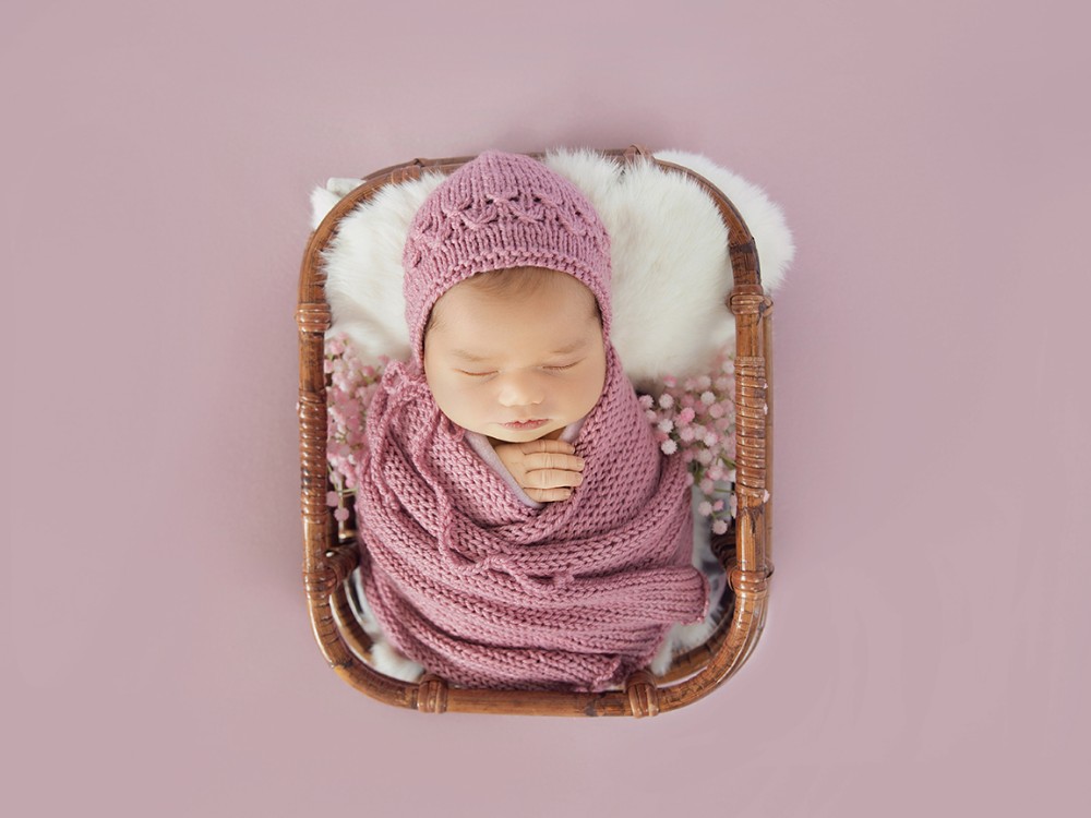 Sleeping newborn baby wrapped in soft pink inside a wooden basket, styled for a bespoke fine art newborn photography session in Witham, Essex
