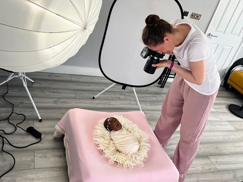 Behind the scenes of a professional mobile newborn photographer creating a safely posed baby portrait with studio lighting inside a client's home in Essex