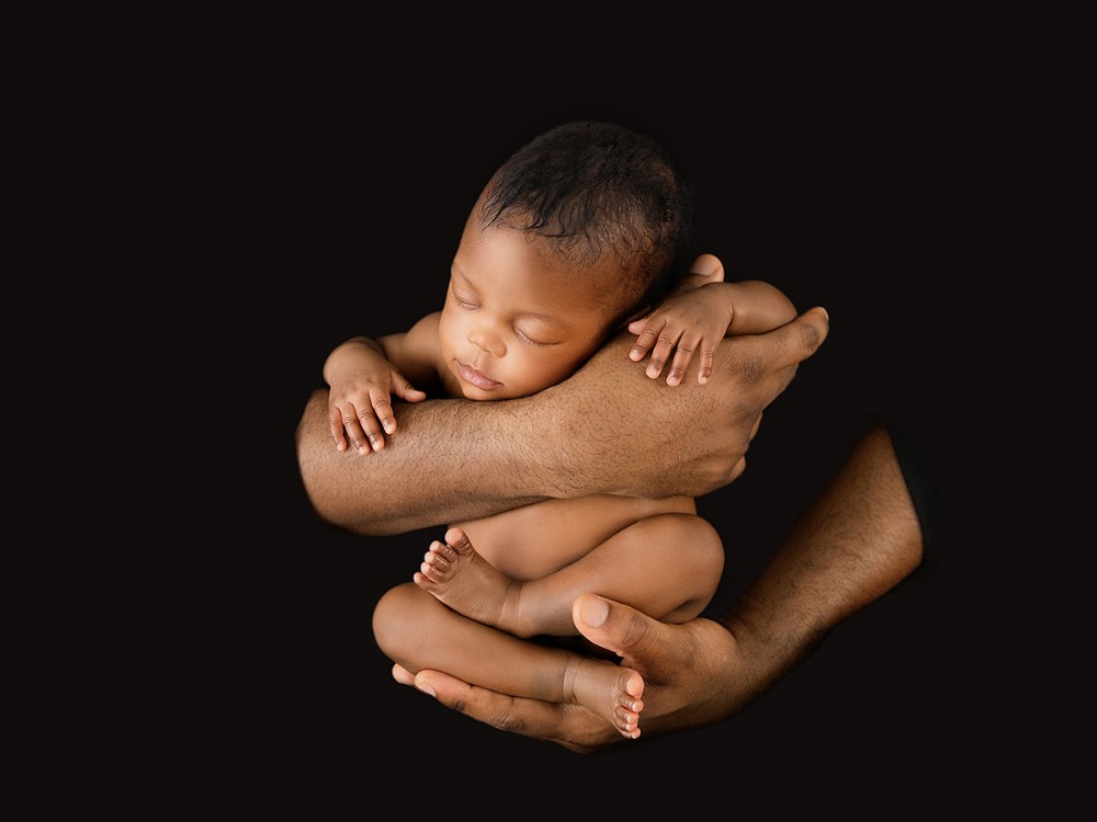 Newborn baby resting safely in father's hands against a dark background, capturing connection during a fine art newborn photography session near Colchester, Essex.