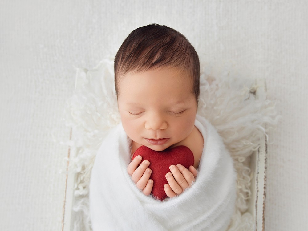 Newborn baby peacefully holding a small red heart in a timeless fine art portrait by an Essex newborn photographer serving Wickford and surrounding areas.