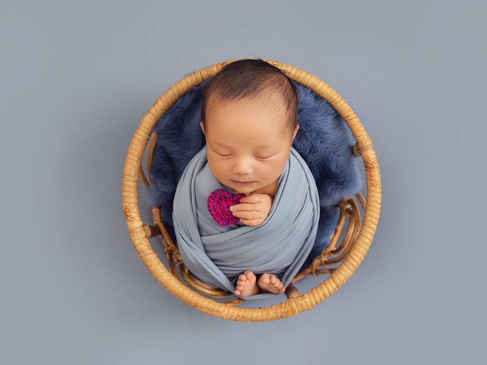 Sleeping newborn baby wrapped in soft blue and posed safely in a natural wooden basket during a fine art newborn photography session near Chelmsford, Essex.