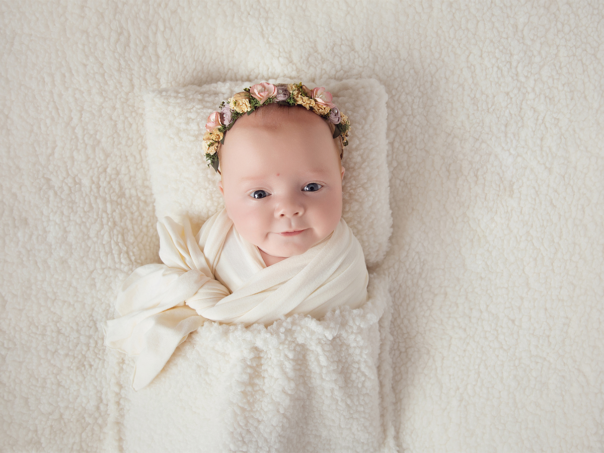 Beautiful newborn baby girl wrapped in soft cream fabric and floral headband during peaceful newborn photography session in Chelmsford Essex.