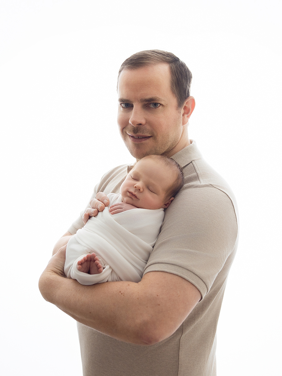 Father wearing a neutral polo shirt while holding his sleeping newborn during an in-home photography session.