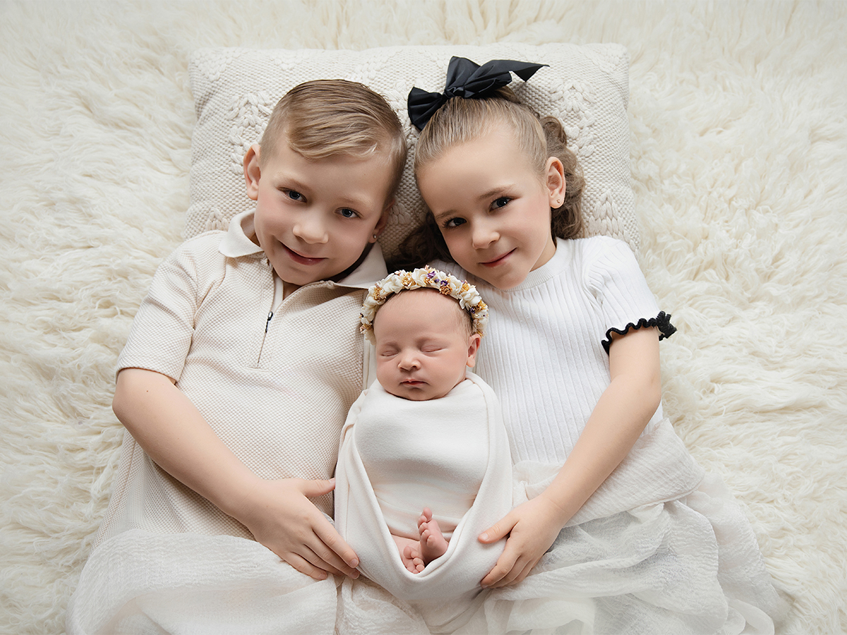 Two siblings lying beside newborn baby in soft neutral setting during sibling newborn photography session in Braintree Essex.