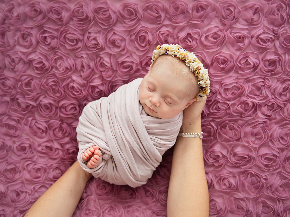 Sleeping newborn baby girl wrapped in soft fabric with a delicate floral headband, photographed on a rose backdrop during a newborn photoshoot in Billericay Essex.