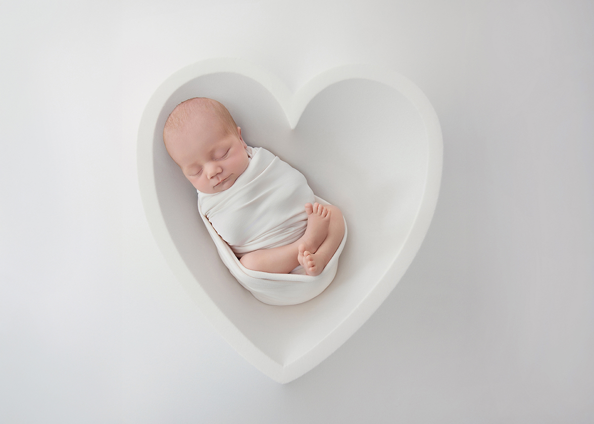 Sleeping newborn baby wrapped in soft fabric posed inside a heart shaped prop during a fine art newborn photography session in Rayleigh Essex.