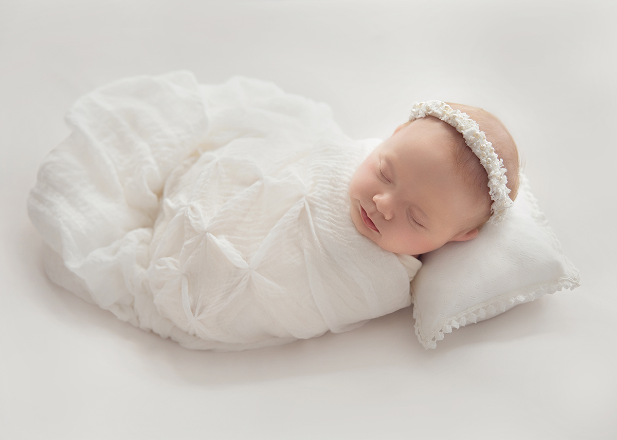 Sleeping newborn baby wrapped in soft white fabric on delicate pillow photographed during newborn photography session in Basildon Essex.