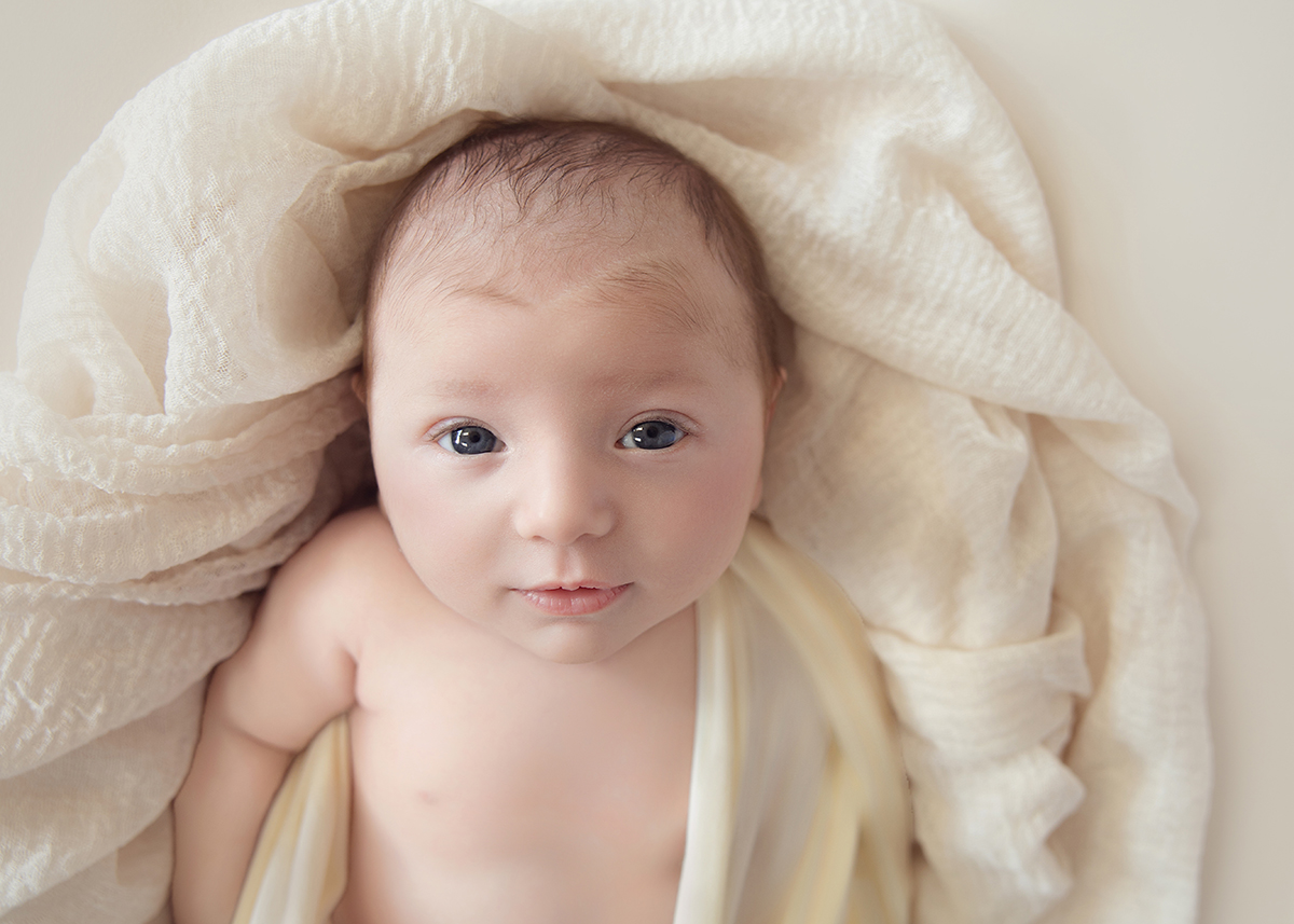Close-up portrait of a wide-eyed newborn baby wrapped in soft cream fabric during a newborn photography session in Basildon Essex.