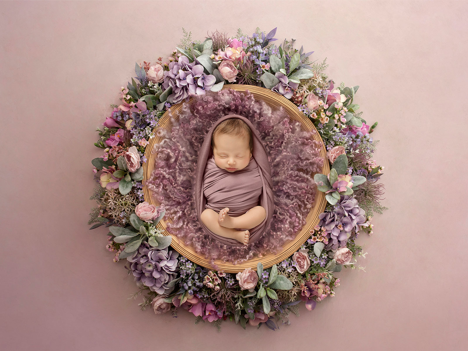 Newborn baby girl peacefully posed in a floral wreath with soft lilac and pink tones during a luxury newborn photoshoot in Essex.